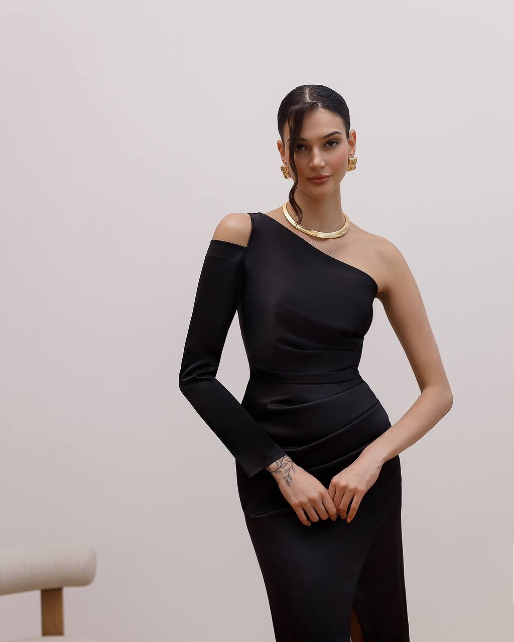 Woman wearing a black one-shoulder dress with gold jewelry against a plain background