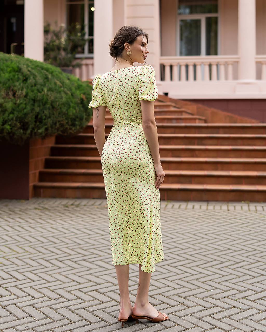 Woman in a light green floral dress standing on a paved area with steps and greenery in the background.