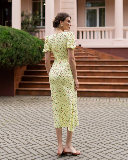 Woman in a light green floral dress standing on a paved area with steps and greenery in the background.