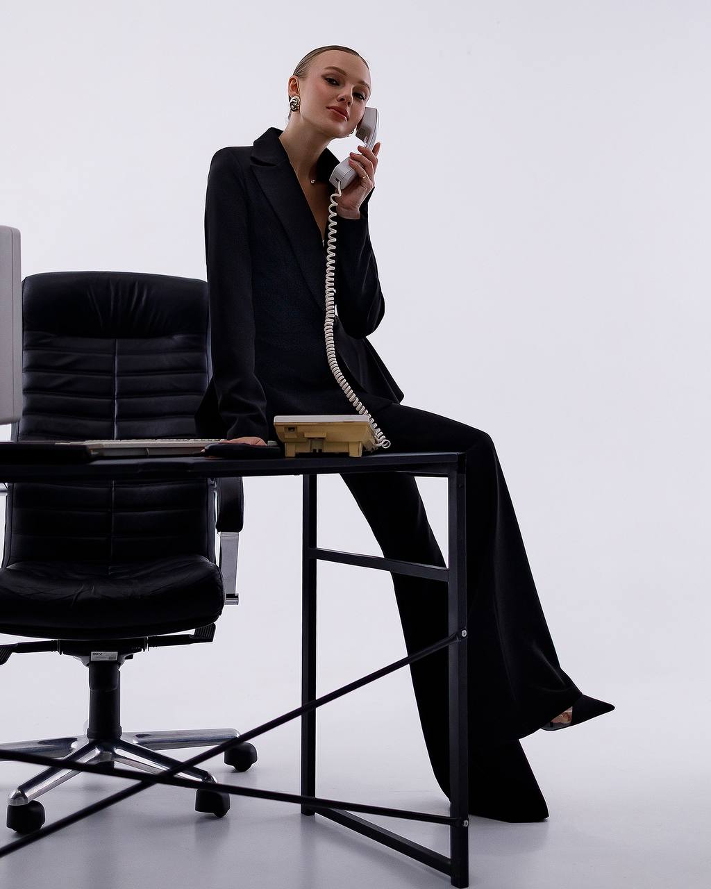 Woman in a black suit sitting on a desk with a phone, in an office setting.
