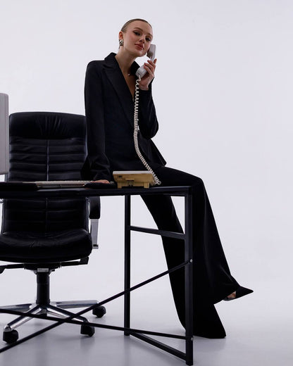 Woman in a black suit sitting on a desk with a phone, in an office setting.