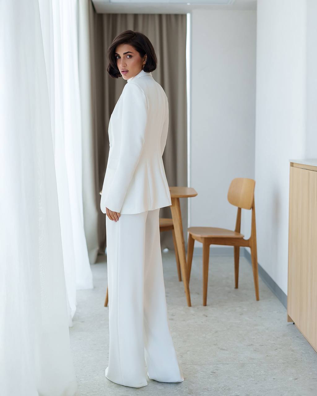 Woman in a white outfit standing in a minimalistic room with wooden furniture.