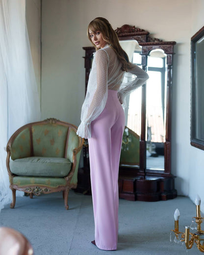 Woman in a white sheer top and pink pants standing in a room with vintage furniture.