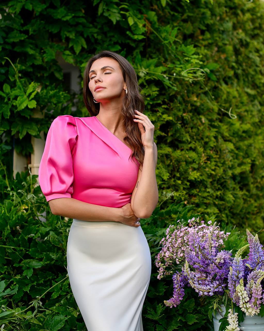 Woman in a pink top and white skirt standing in front of green foliage with flowers.