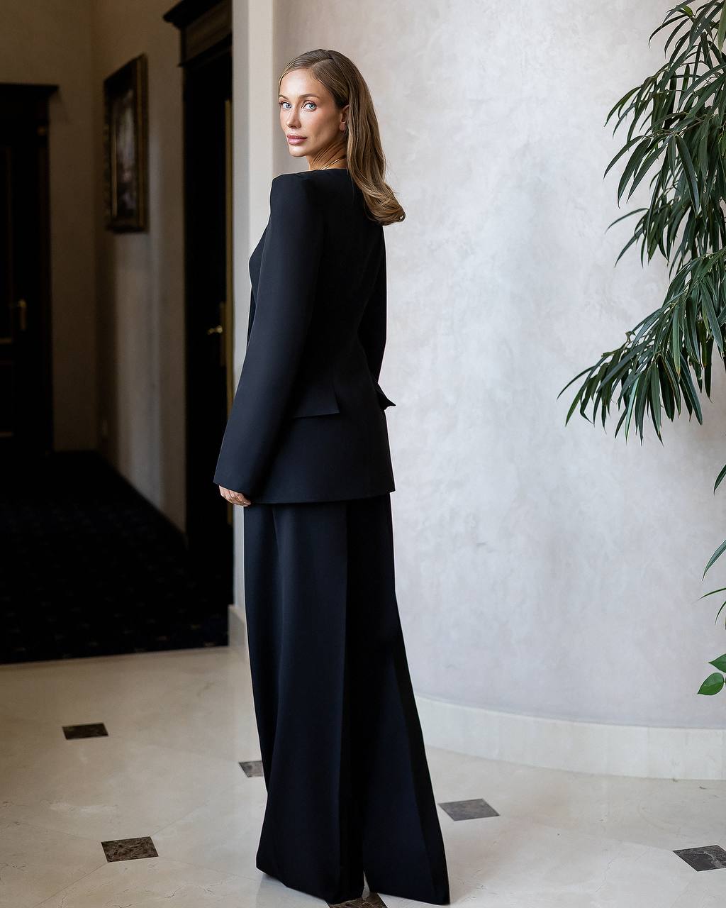 Woman in a black suit standing in an indoor setting with a plant and door in the background.