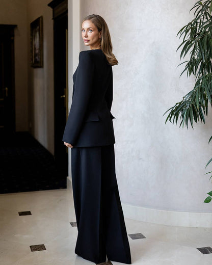 Woman in a black suit standing in an indoor setting with a plant and door in the background.