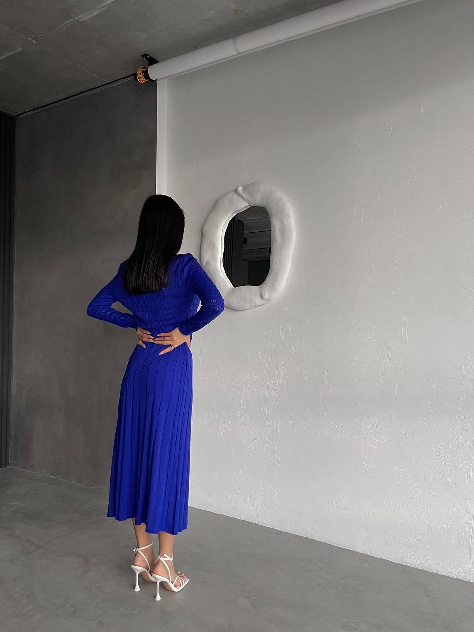 Woman in a blue dress standing in a minimalistic room with a white wall and circular mirror.