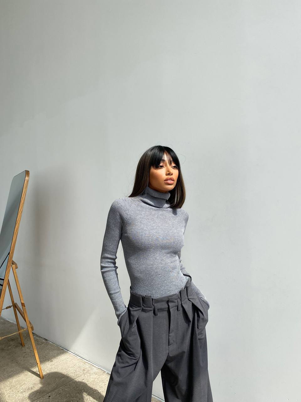 Person wearing a gray turtleneck and dark pants against a plain wall.