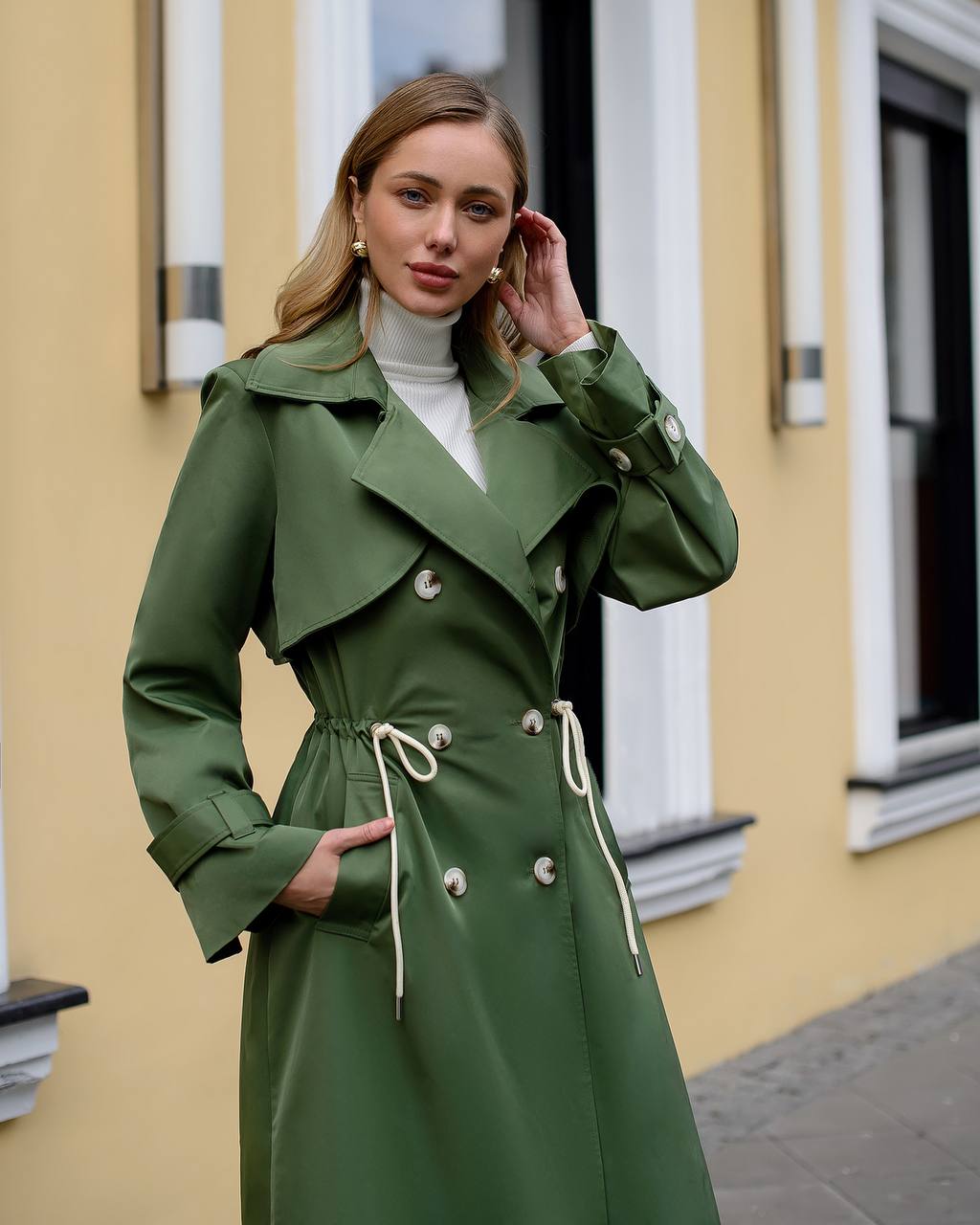 Woman wearing a green trench coat standing in front of a yellow building.