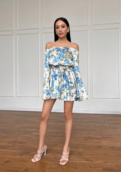Woman wearing a floral off-shoulder dress standing against a white paneled wall.