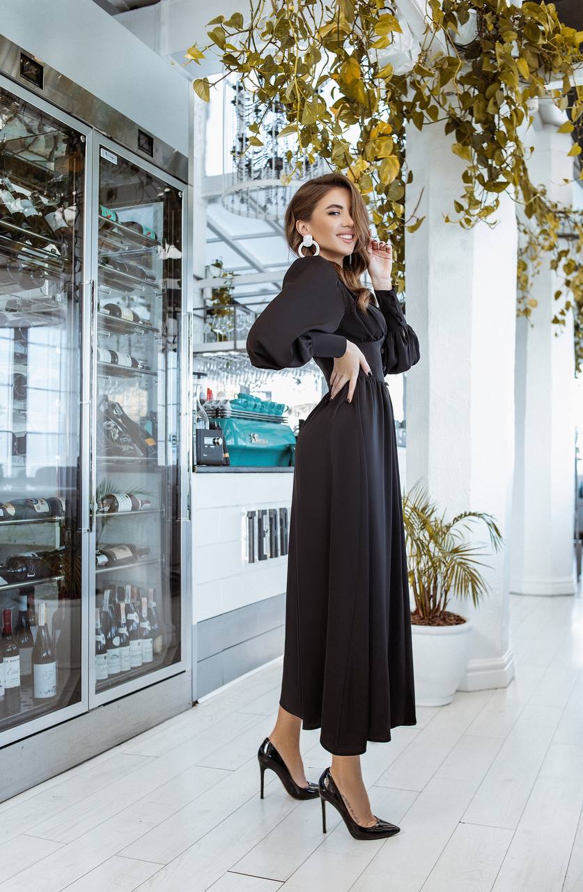 Woman in a black dress standing in a modern interior setting with plants and glass doors.