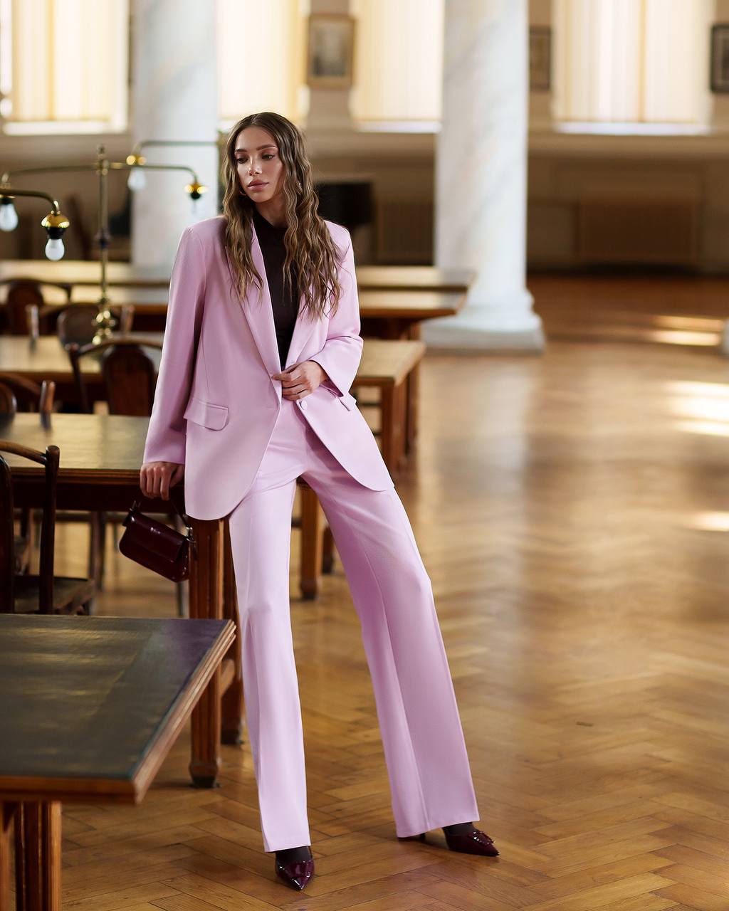 Woman in a pink suit standing in an indoor setting with wooden floors and tables.