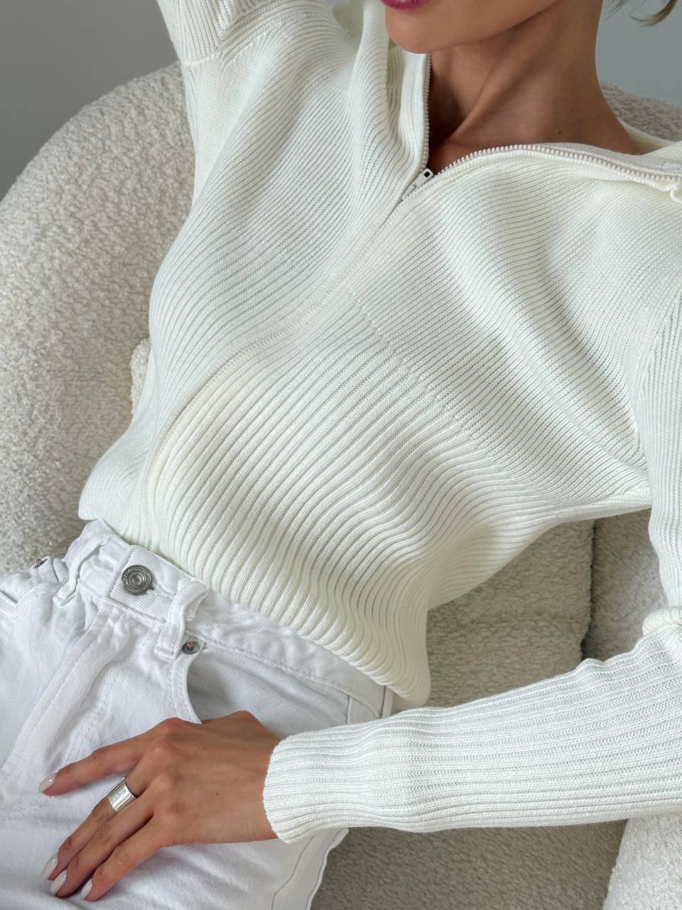 Person wearing a white ribbed sweater and white pants sitting on a beige chair.