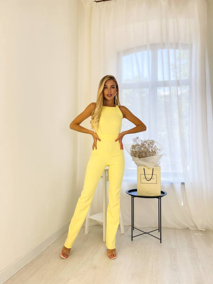 Woman in a yellow jumpsuit posing in a room with white curtains and a small table with a plant.
