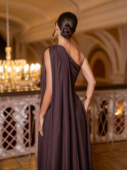 Woman in a dark brown one-shoulder dress standing in an elegant indoor setting with chandeliers.