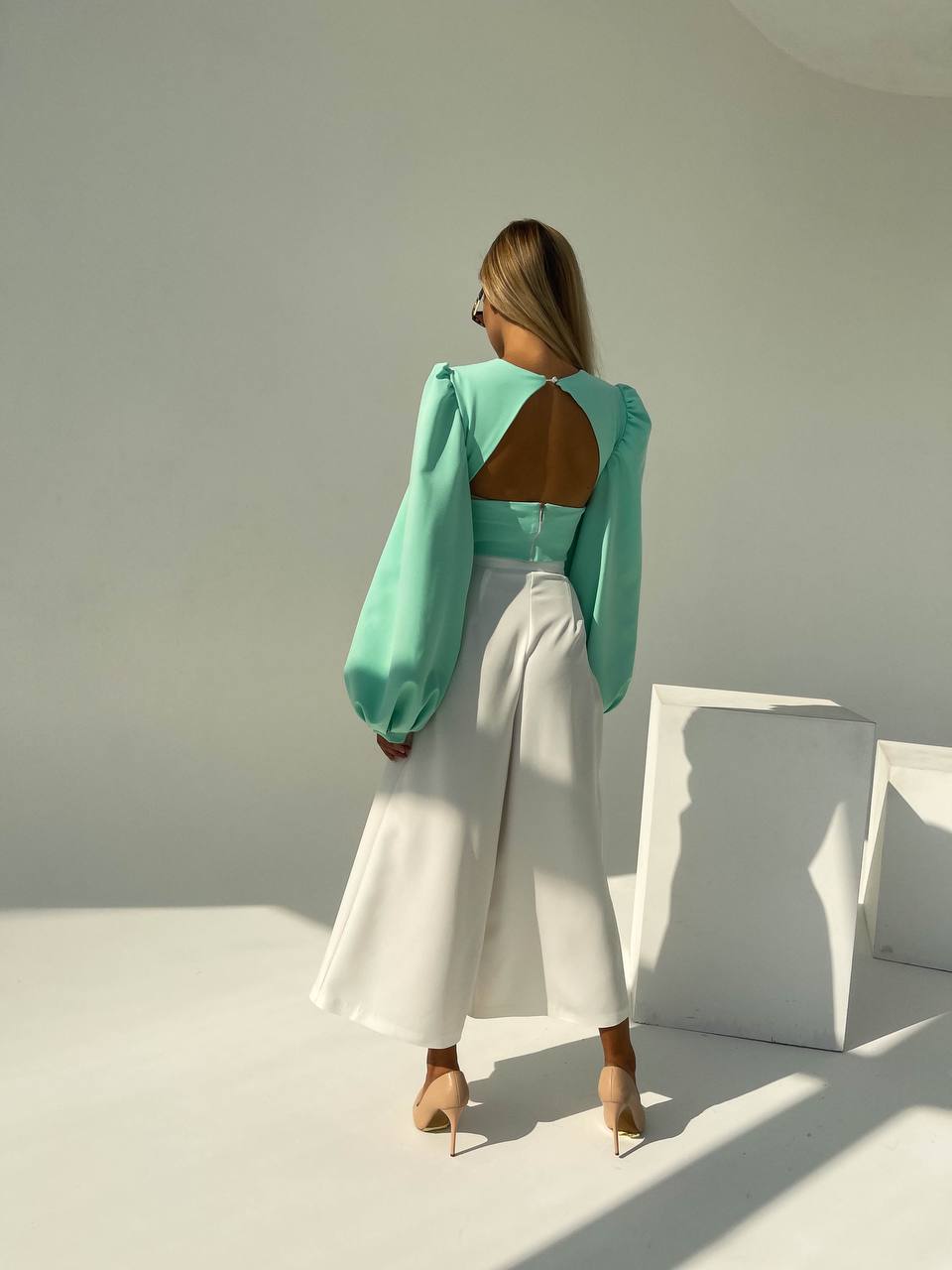 Woman wearing a turquoise top with cut-out design and white pants in a minimalistic setting.