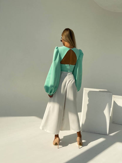 Woman wearing a turquoise top with cut-out design and white pants in a minimalistic setting.