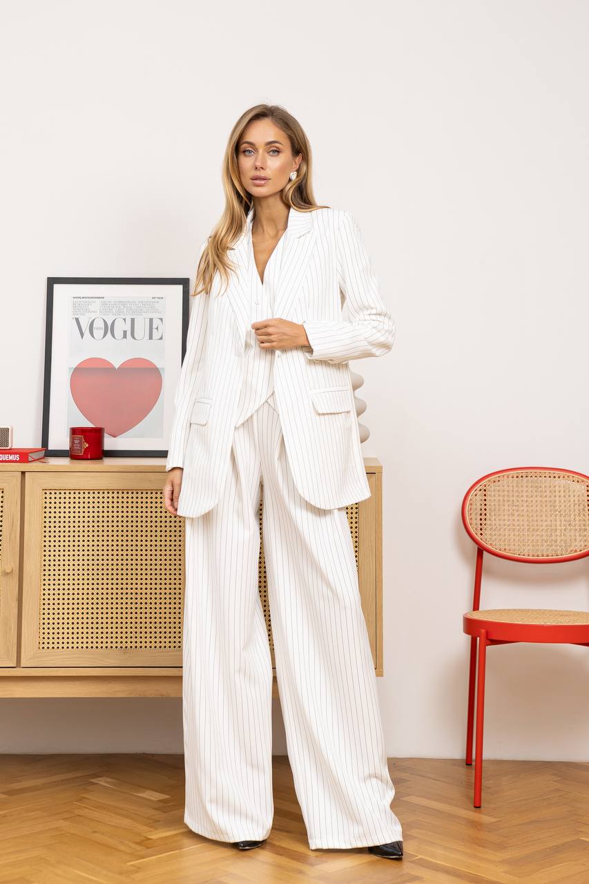 Woman in a white suit standing in a room with a red chair and framed Vogue magazine.