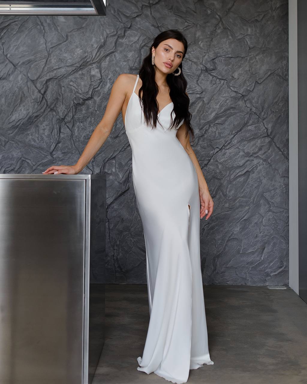Woman in a white dress standing against a textured gray wall.