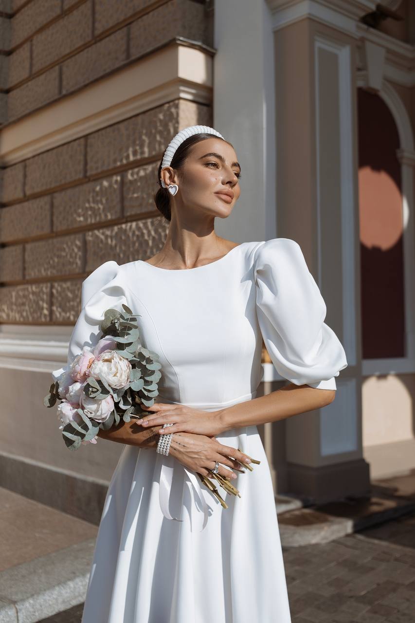 Woman in a white dress holding a bouquet of flowers, standing in front of a building.