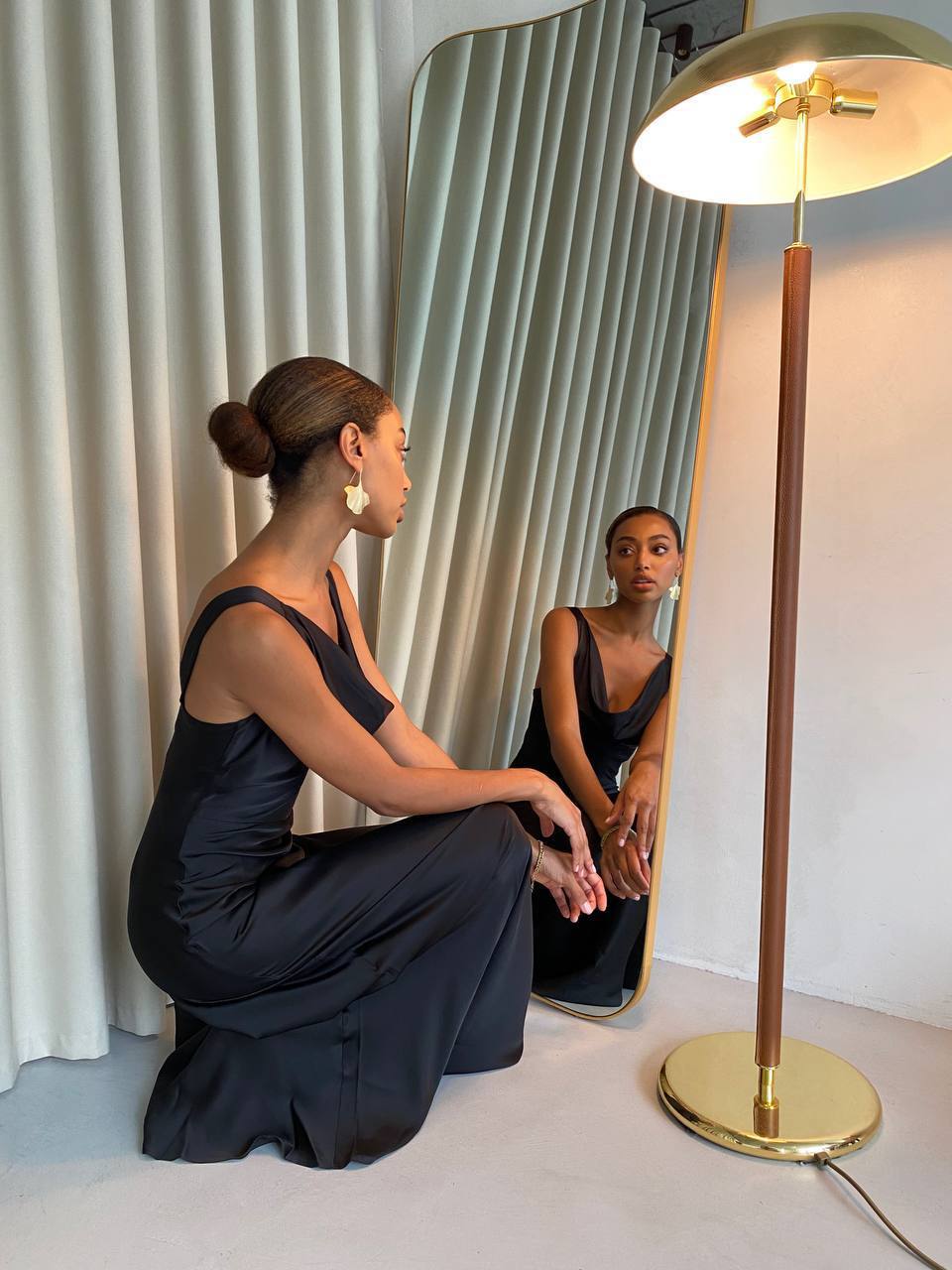 Woman in a black dress sitting on the floor in front of a mirror, with a gold lamp beside her.