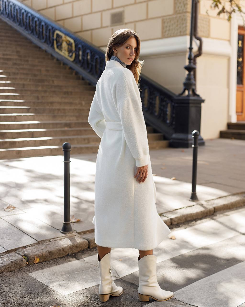 Woman in a white coat standing on a city street with steps and a building in the background.