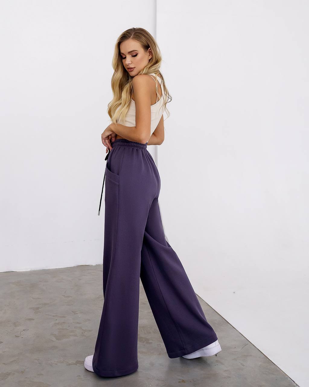 Woman wearing a white top and purple wide-leg pants against a white wall.