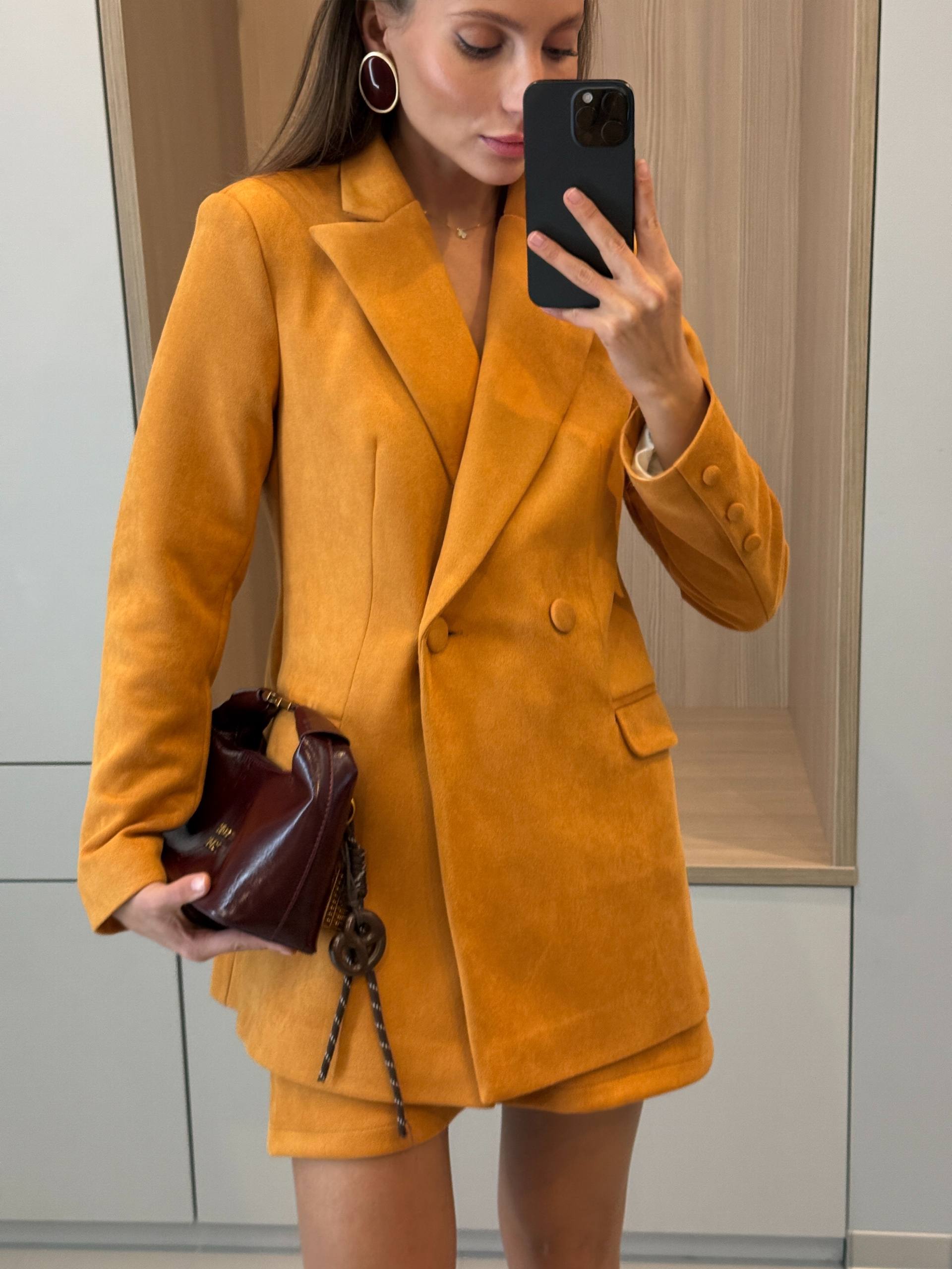 A woman takes a mirror selfie wearing a mustard yellow double-breasted blazer and matching shorts, holding a dark brown leather handbag. The background shows a modern interior with light wood paneling and white cabinets.
