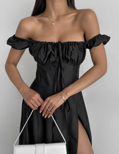 Woman wearing a black off-shoulder dress with a white handbag on a plain background