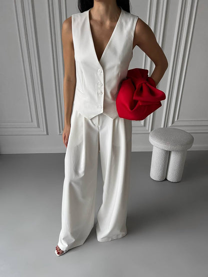 Person wearing a white sleeveless top and pants holding a red bag in a minimalistic setting.