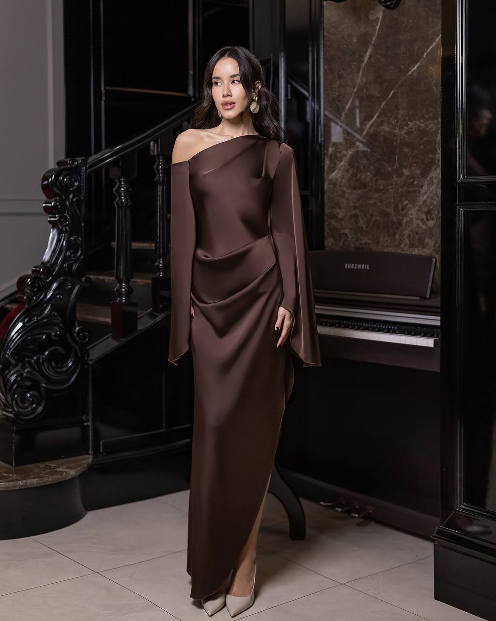Woman in a brown off-shoulder dress standing in an elegant interior setting.