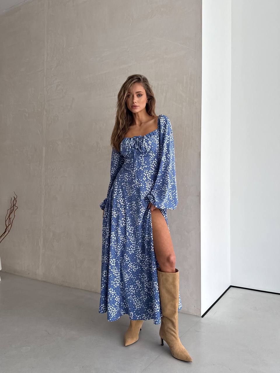 Woman wearing a blue floral dress with a slit, standing against a neutral wall.