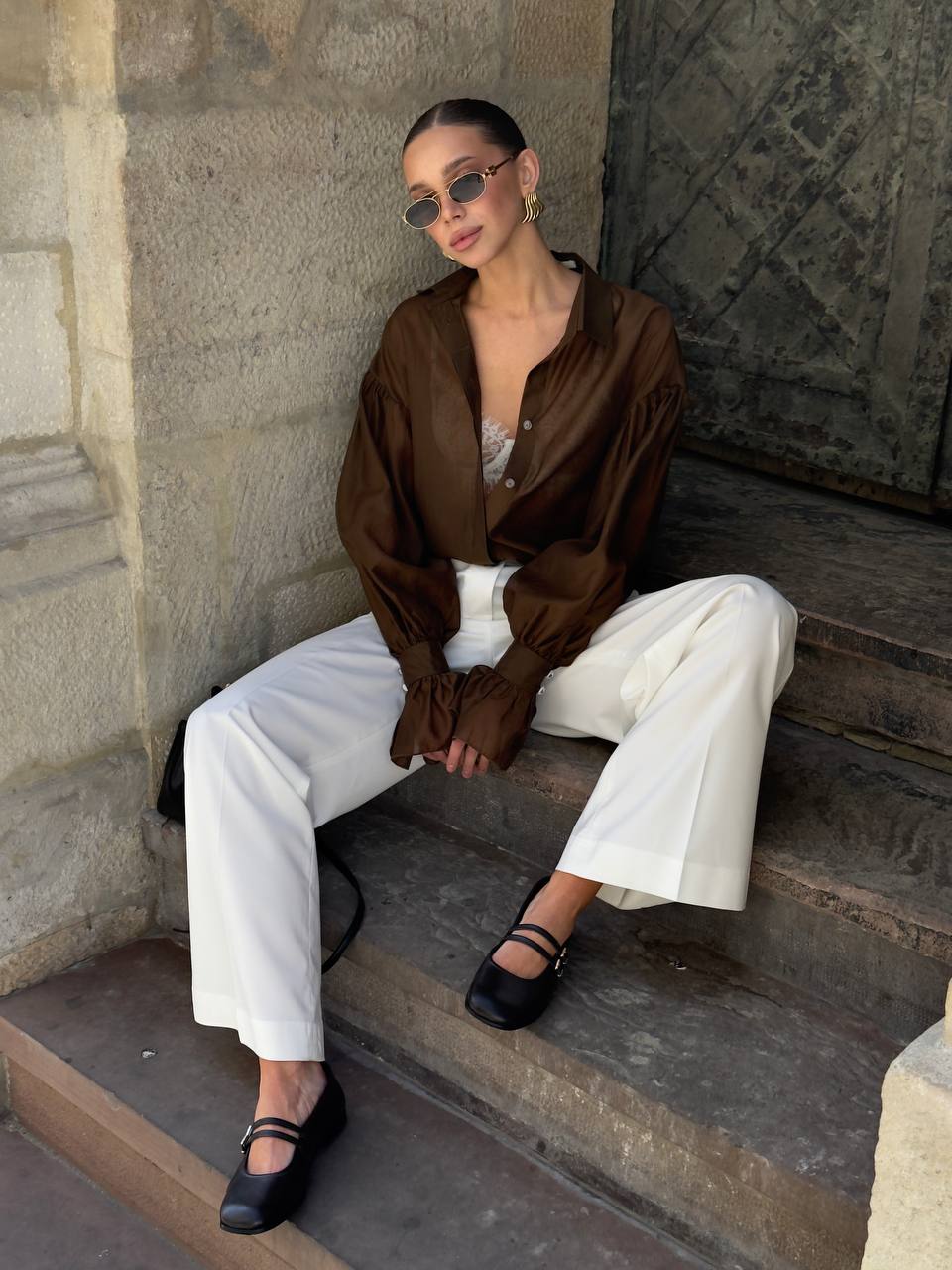 Woman sitting on stone steps wearing a brown blouse, white pants, and black shoes.