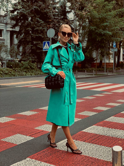 Woman in a turquoise trench coat crossing a street with a black handbag.