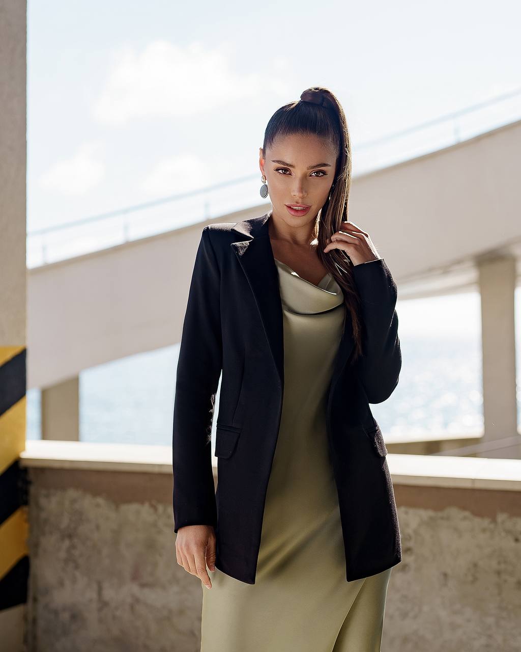 Woman in a black blazer over a green dress standing indoors with large windows.
