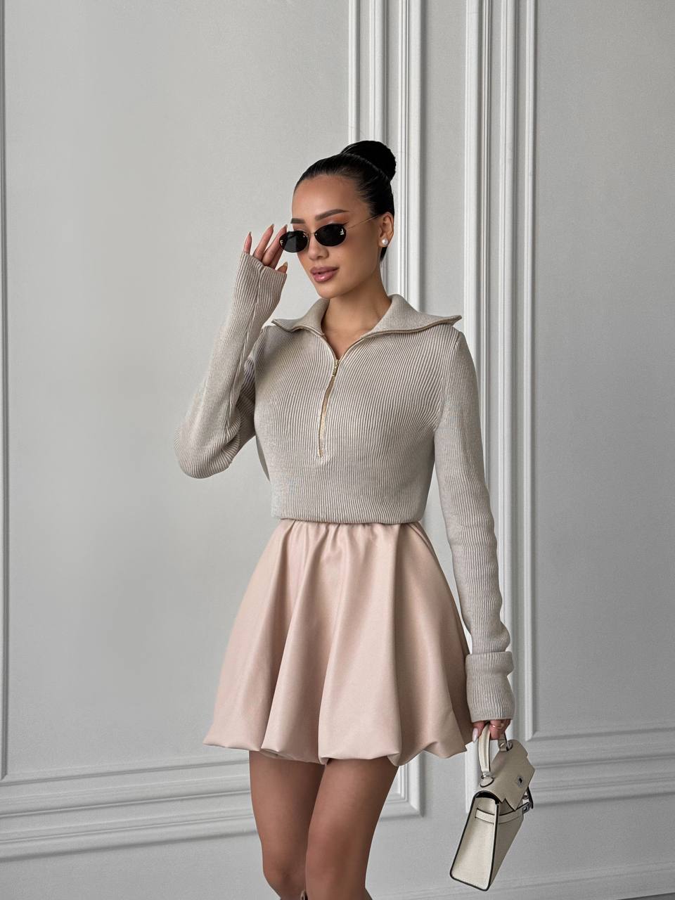 Woman wearing a beige sweater and pink skirt against a gray wall.