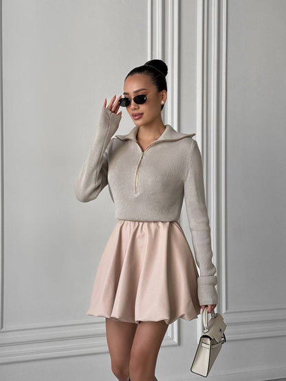 Woman wearing a beige sweater and pink skirt against a gray wall.