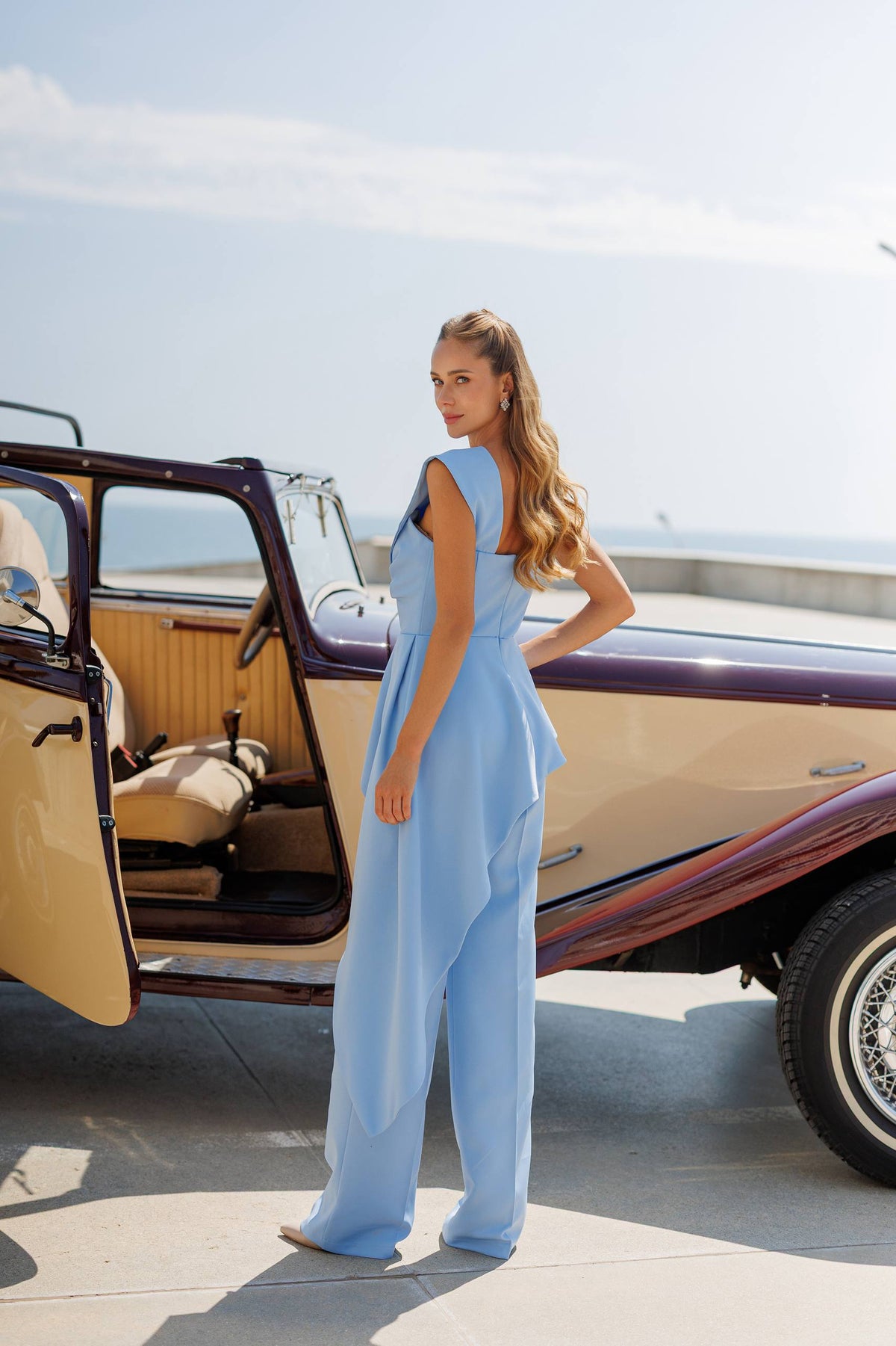 Woman in a light blue dress standing next to an open car door on a sunny day.