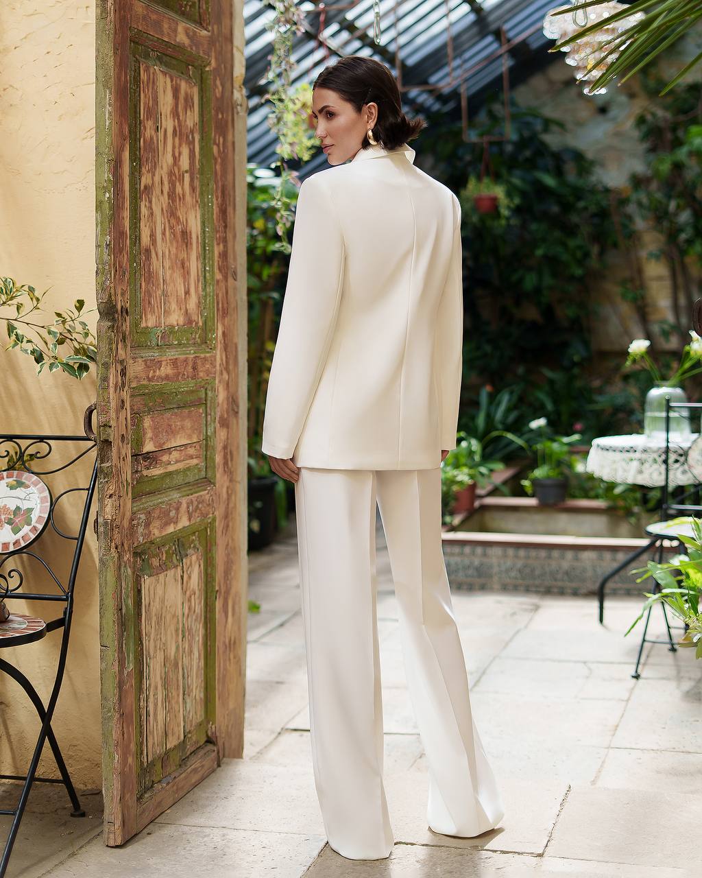 Woman in a white suit standing in a garden setting with a rustic door and plants.