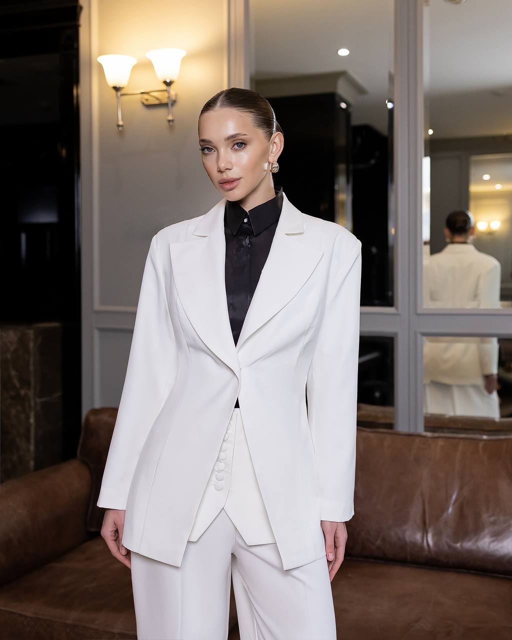 Woman in a white suit standing in a room with a mirror and lamp.
