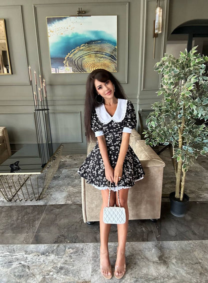 Woman in a black floral dress with a white collar holding a white handbag in a stylish interior setting.