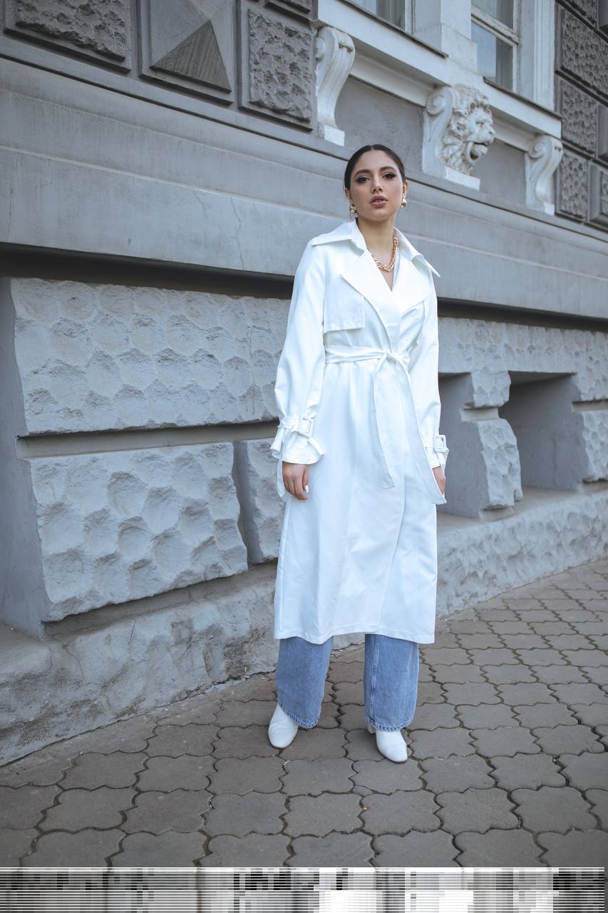 Woman in a white coat and blue pants standing in front of a stone building.