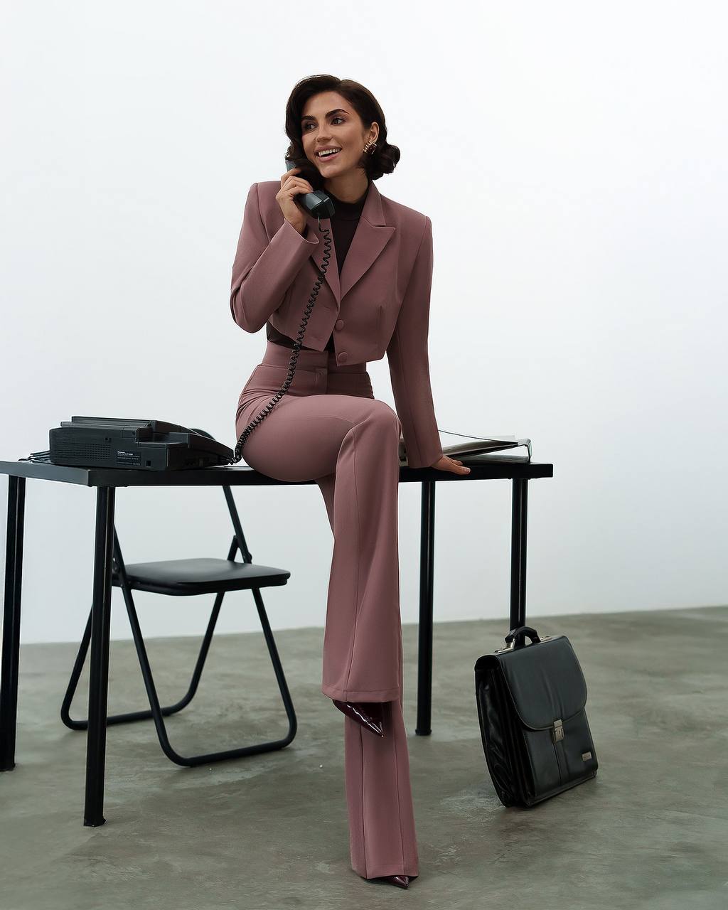 Woman in a pink suit sitting on a black stool with a black bag beside her.