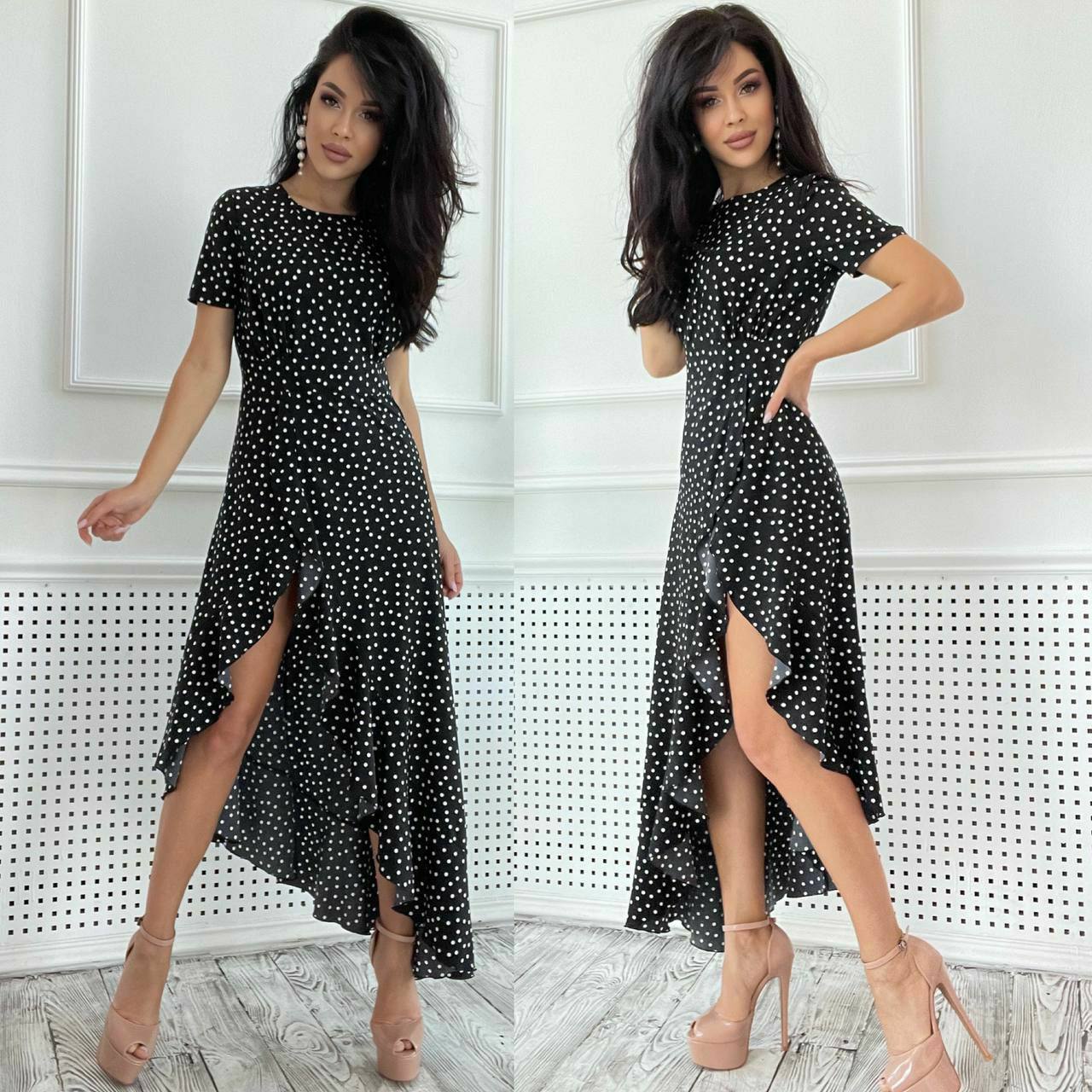 Woman wearing a black polka dot dress with a high-low hem in a white room.