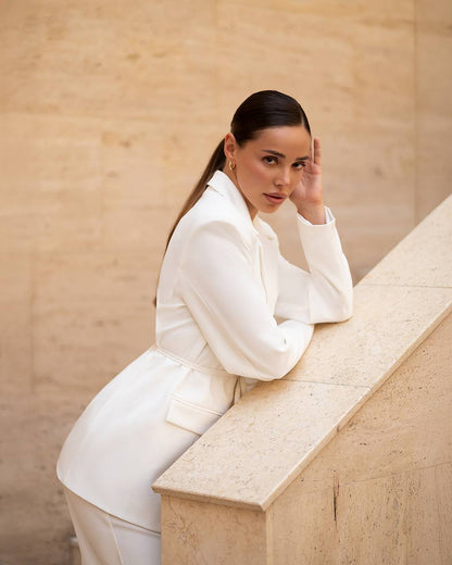 Woman in a white outfit sitting on a stone ledge against a beige wall.