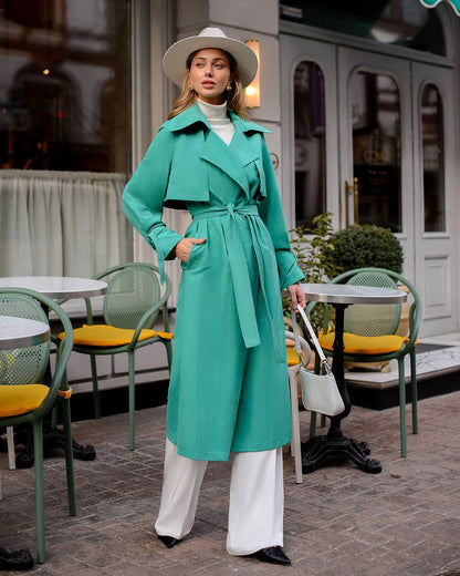 Woman in a teal trench coat standing in an outdoor cafe setting