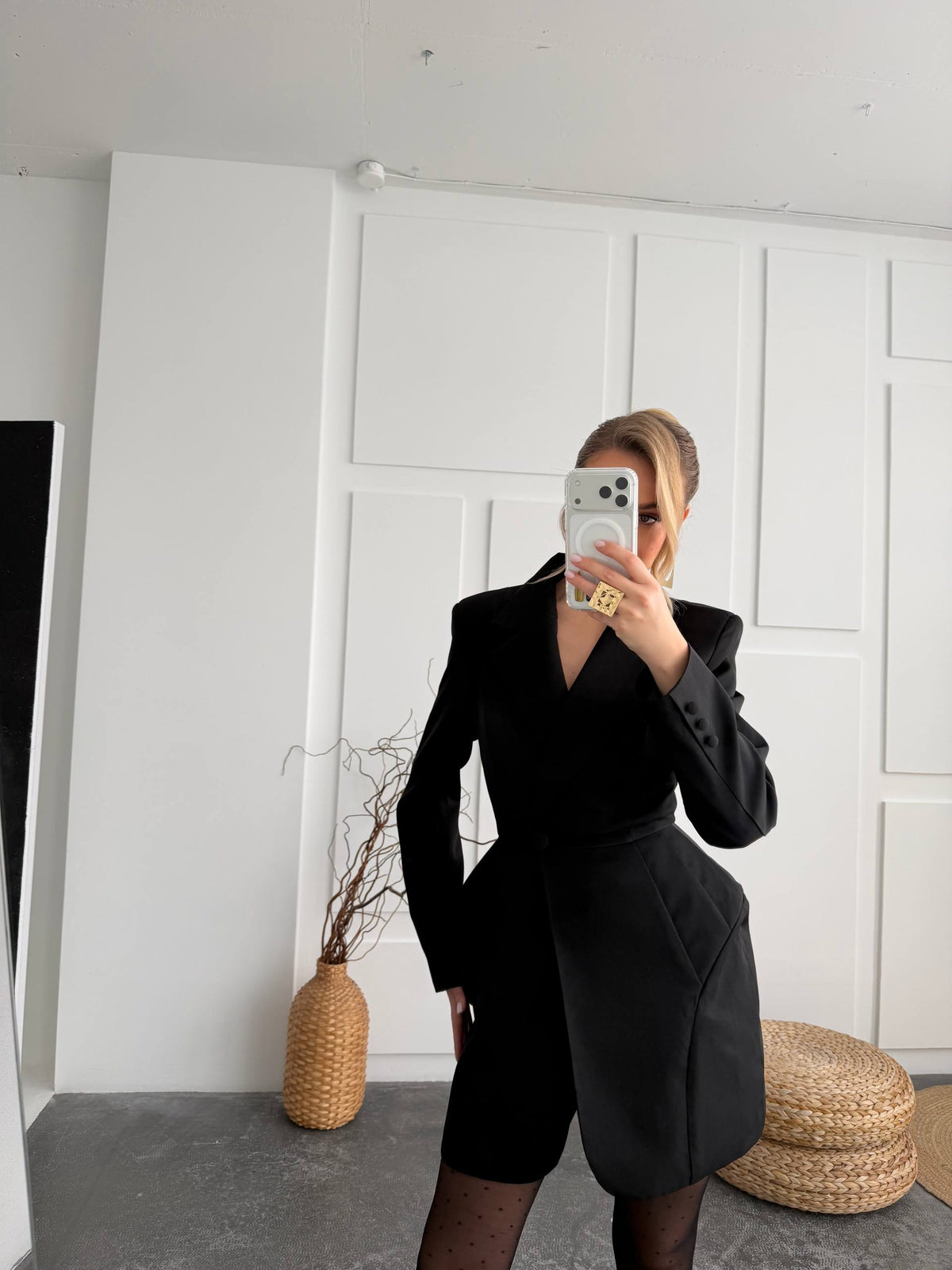 A woman takes a mirror selfie wearing a black structured blazer-style dress with a deep V-neck and asymmetrical wrap-style skirt, paired with sheer black tights. The setting is a minimalist room with white paneled walls, a woven vase, and a round woven pouf.