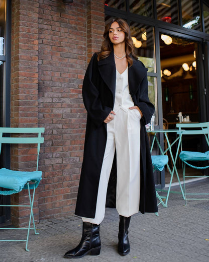 Woman in a black coat and white pants standing outdoors near a brick wall.
