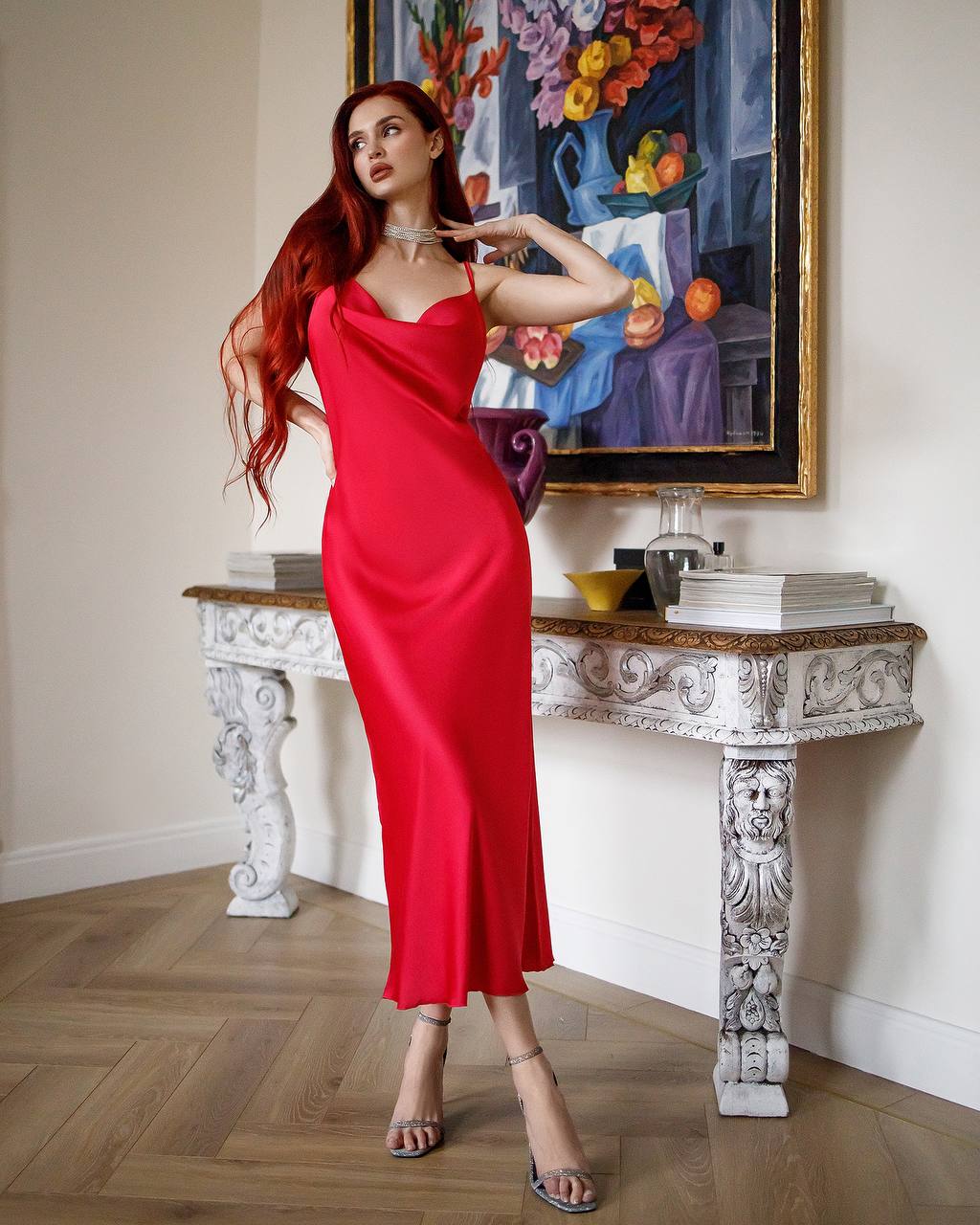 Woman in a red dress posing in an elegant room with a decorative table and artwork.