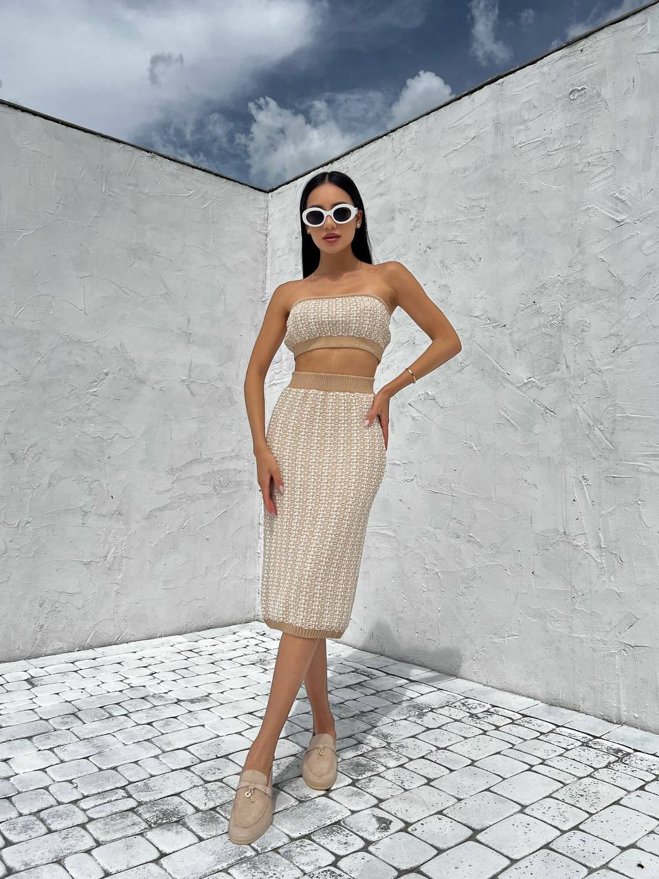 Woman in a beige outfit standing in front of a white wall with a blue sky.